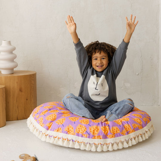 Child sitting on a colourful round cushion with raised arms, wearing a grey sweatshirt