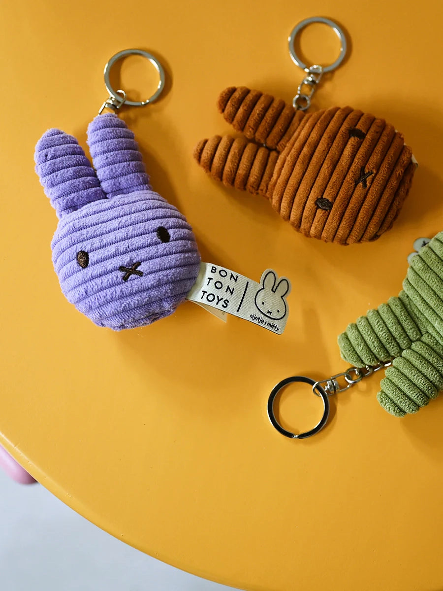 Three colourful keyrings, one cinammon brown, one lilac and one sage green on a yellow surface with a visible brand label. All are made with chunky corduroy material
