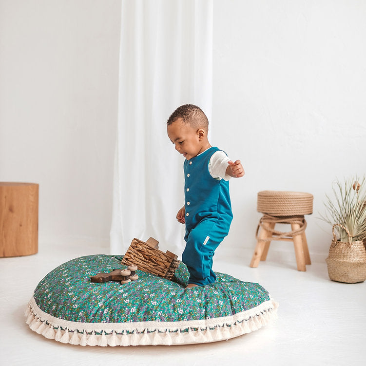 Child playing on a round green cushion with floral pattern in a room with white curtains and wooden furniture.