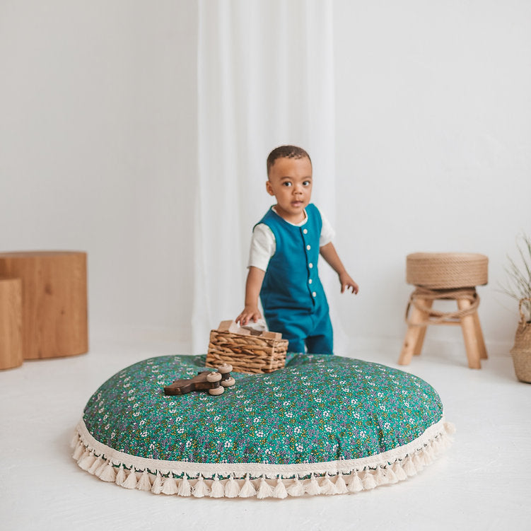 Child standing on a floral cushion in a minimalistic room with wooden blocks and a stool.
