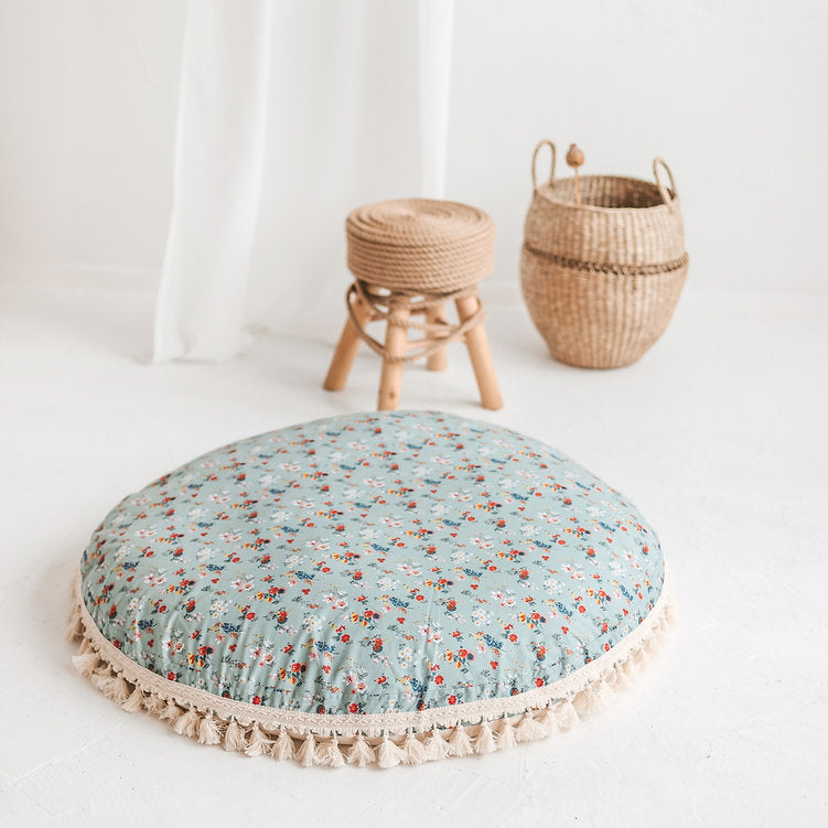 Round floral cushion with ruffle trim on a white floor, with a small stool and basket in the background.