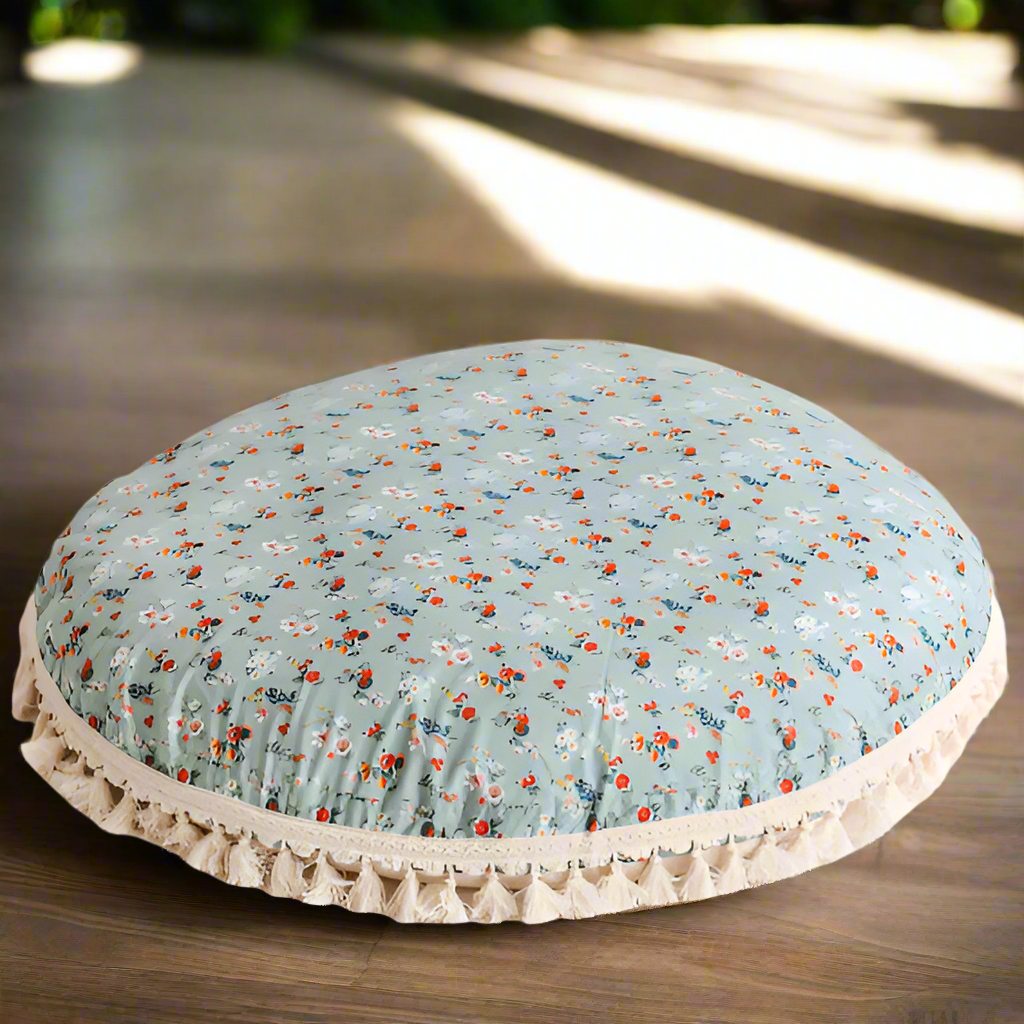 Round floor cushion with a light blue/mint background  and a floral pattern with white, red and blue flowers, and white tassels around the edge