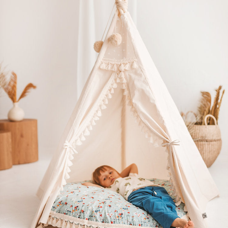 Child lying on a floral floor cushion inside a white teepee with decorative elements in a room.