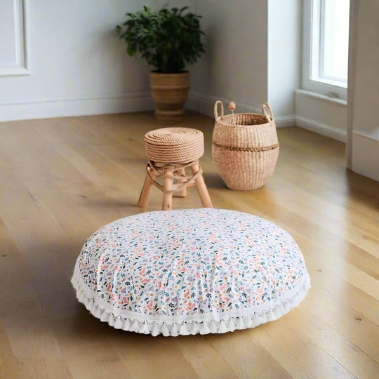 Round floral-patterned cushion with tassels on a white floor, with a small wooden stool and woven basket in the background.