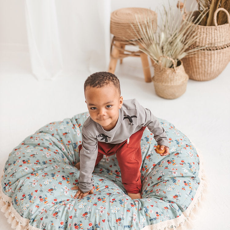 Child standing on a floral cushion in a home setting with wicker baskets and plants.