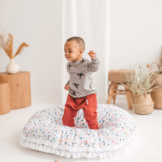 Child standing on a floral cushion in a room with wooden furniture and decorative plants.