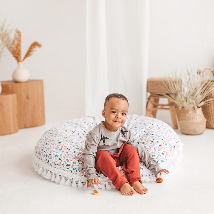 Child sitting on a round cushion in a room with wooden blocks and decorative plants.