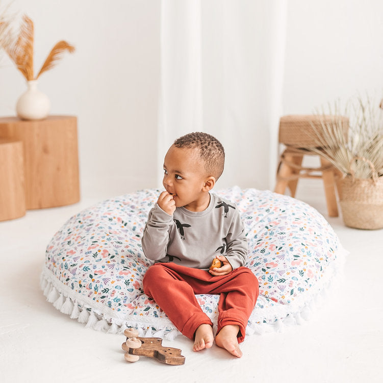 Child sitting on a round cushion in a room with wooden blocks and plants.