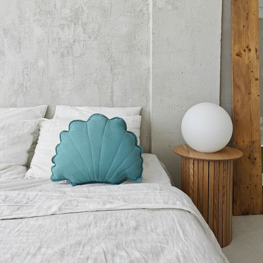 Teal shell-shaped pillow on a bed next to a wooden side table with a white lamp.