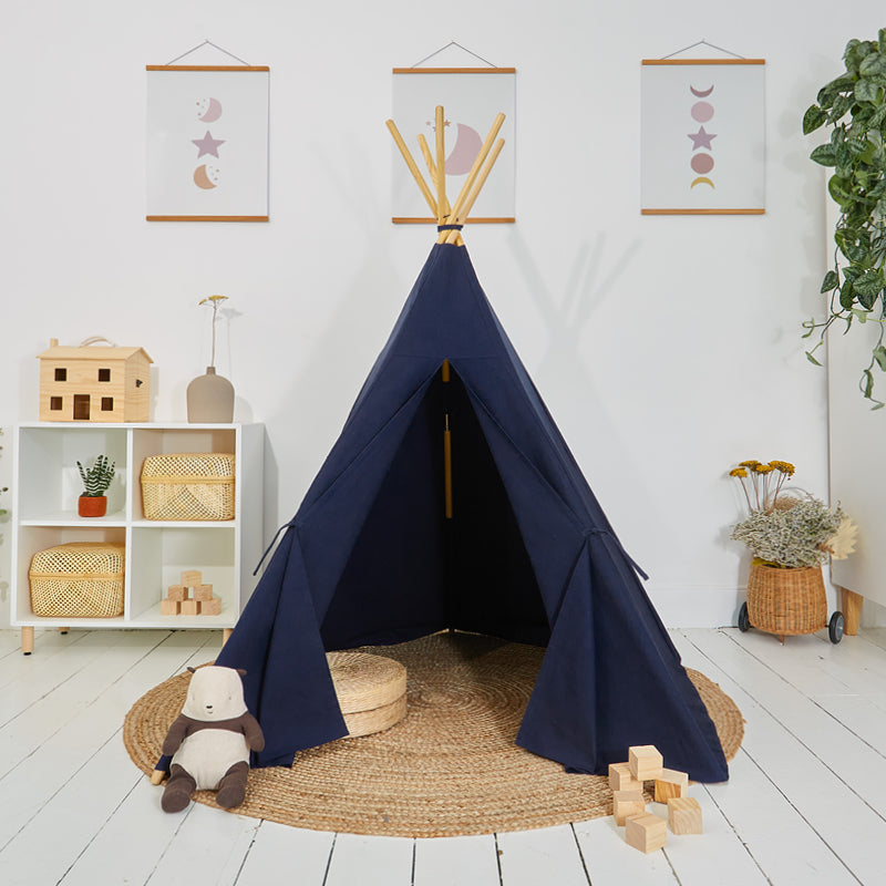 A navy blue play teepee made from cotton canvas, set up on a white room with a wooden five-pole system. Toys and a carry case are visible around the teepee.