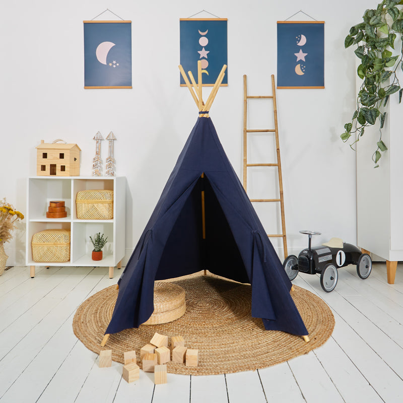 A navy blue play teepee made from cotton canvas, set up on a round jute rug in a room with white flooring. The teepee has a solid wooden five-pole system.