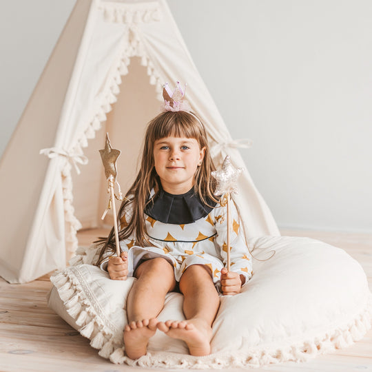Young girl sitting on a white cushion in front of a white teepee, holding fairy wings and a wand.