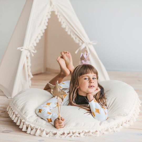 Child lying on a floor cushion in front of a teepee with a crown on, holding a wand.