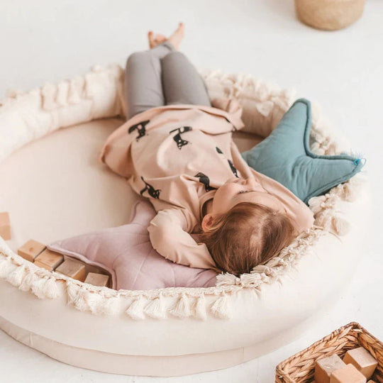 Child lying on a round white bed with toys around, wearing a pink coat with black patterns.