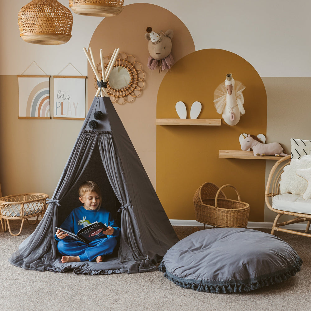 Child sitting inside a grey teepee reading a book in a cosy room with toys and decor.