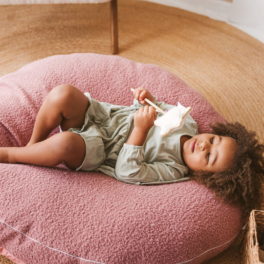 Child lying on a pink cushion holding a toy in a room with wooden floor.