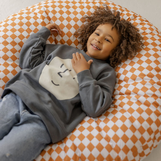Child wearing a grey hoodie with a smiley face design, lying on an orange and white chequered floor cushion