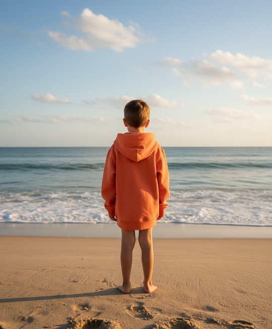 Person wearing an orange hoodie standing on a beach looking at the ocean.