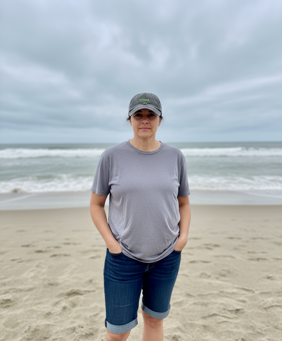 Person standing on a beach wearing a gray t-shirt and cap with a cloudy sky.
