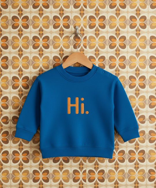 Blue sweatshirt with 'Hi.' embroidered on it hanging on a wooden hanger against a patterned wall.