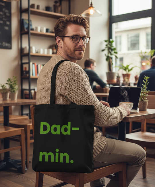 Man sitting in a cafe with a black tote bag that says 'Dad-min' on the back.