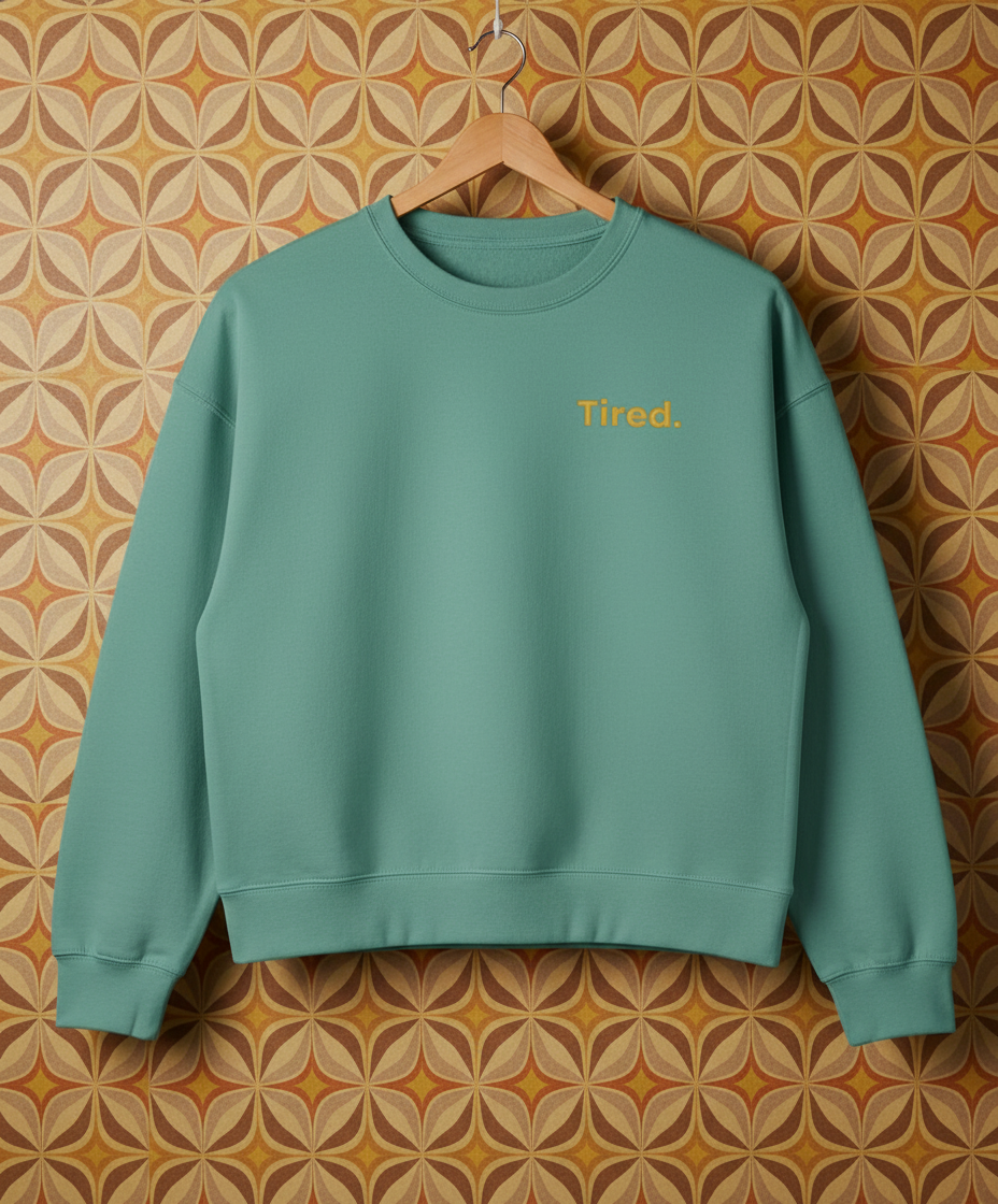 Green sweatshirt with 'Tired.' text on a hanger against a patterned wall.