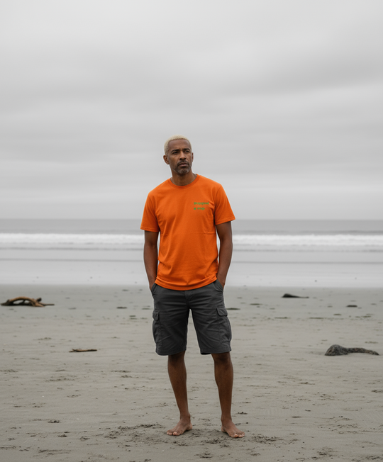 Man wearing an orange shirt and gray shorts standing on a beach with a cloudy sky.