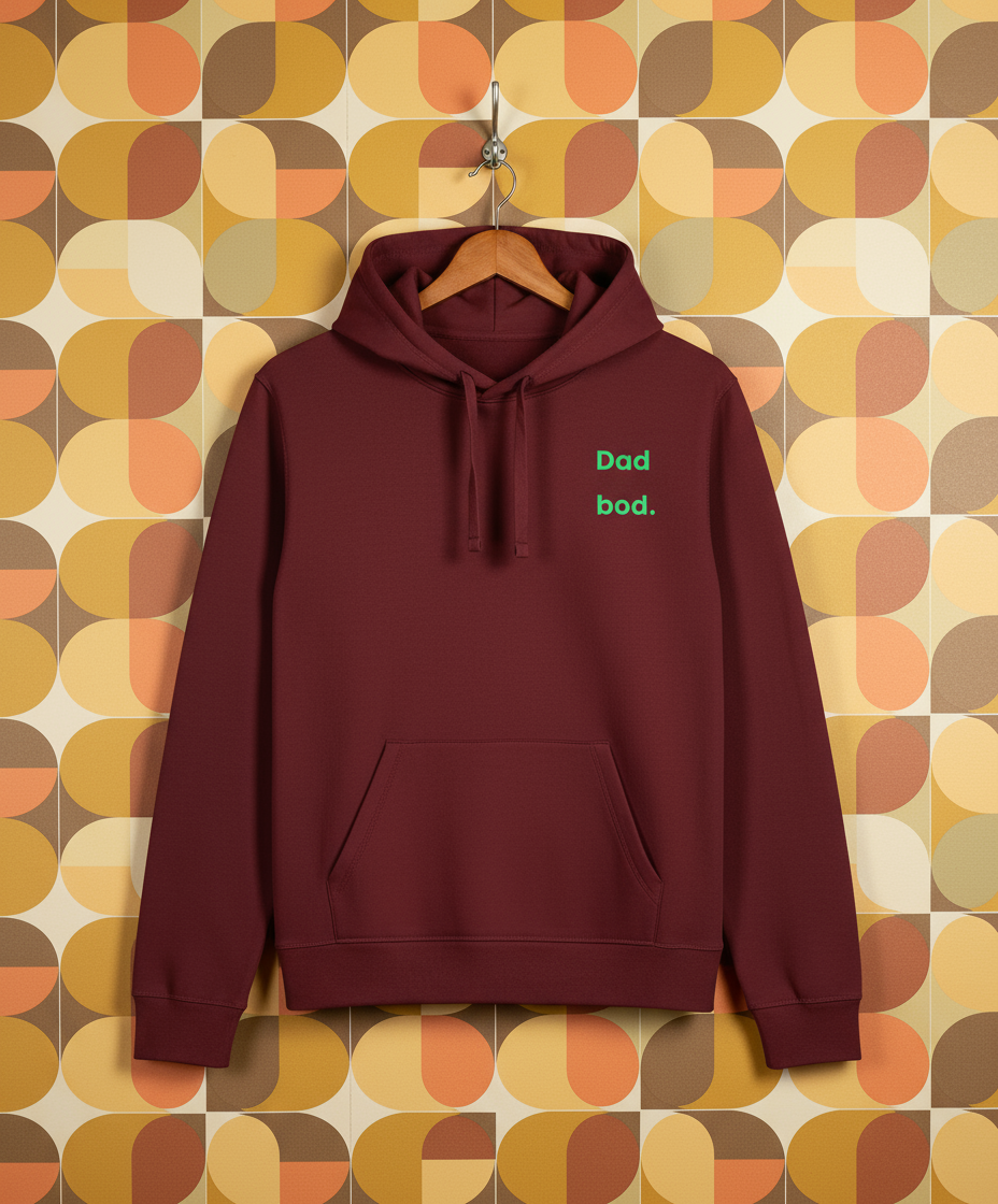 Maroon hoodie with 'Dad bod.' text on a hanger against a patterned wall.