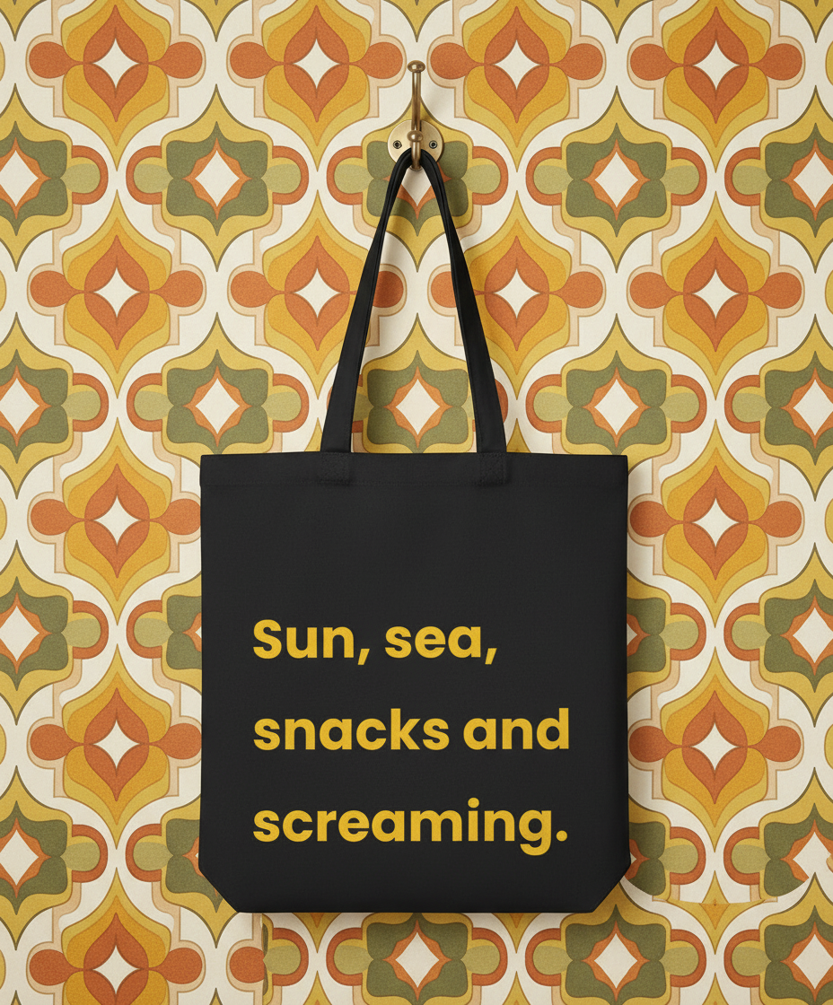 Black tote bag with yellow text 'Sun, sea, snacks and screaming' on a patterned wall background