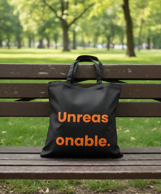 Black tote bag with orange text 'Unreas onable.' on a wooden bench in a park.