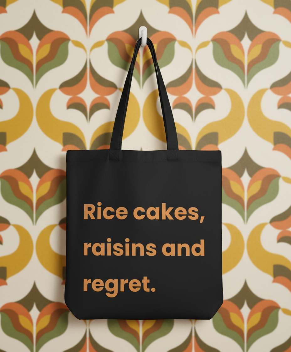 Black tote bag with text 'Rice cakes, raisins and regret' on a patterned wall background