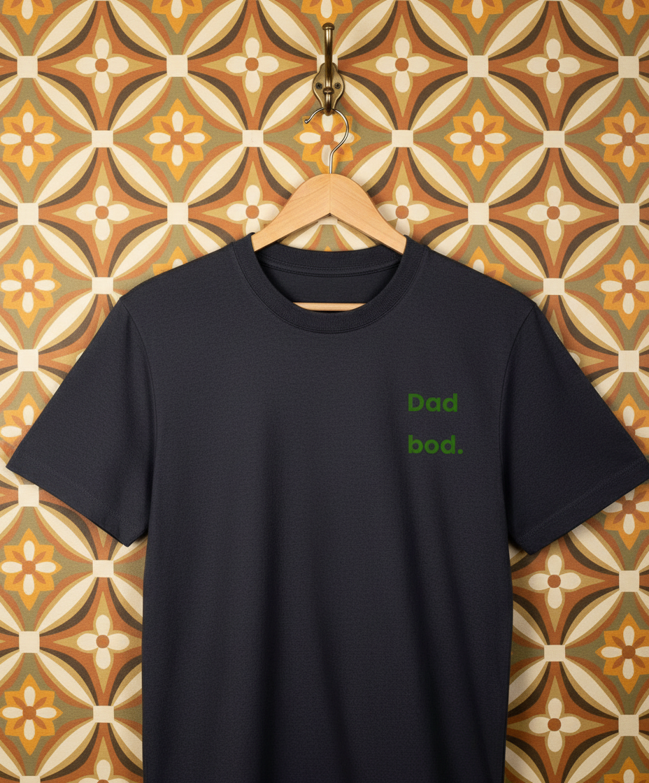 Navy t-shirt with 'Dad bod.' text on a hanger against a patterned wall.