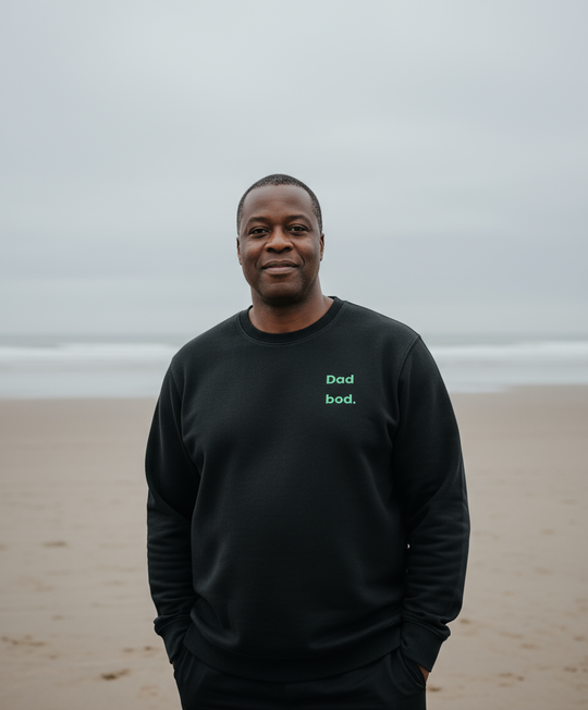 Man wearing a black sweatshirt with 'Dad bod.' text on a beach