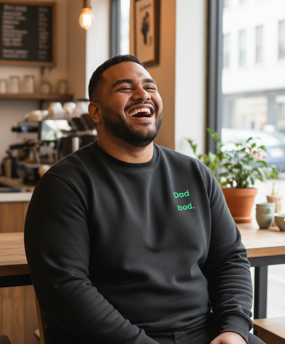 Man laughing in a coffee shop wearing a black sweatshirt with text.