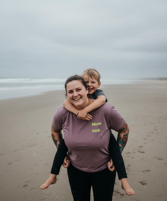 Woman carrying a child on her back at the beach with 'Mum bod.' shirt
