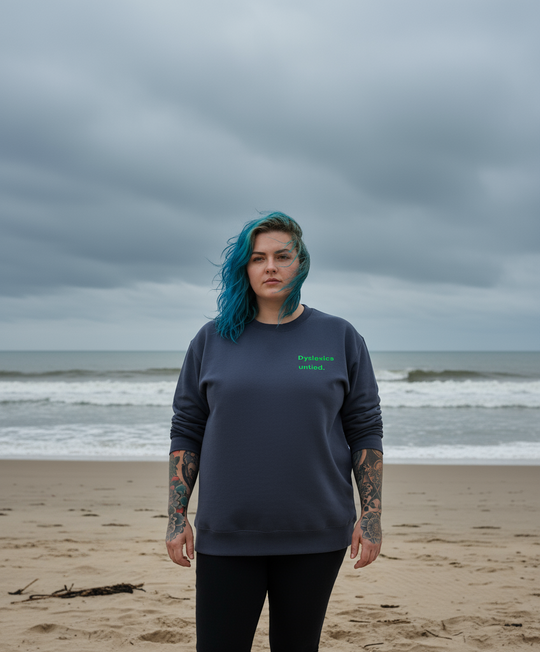 Person with blue hair standing on a beach with a dark grey sweatshirt
