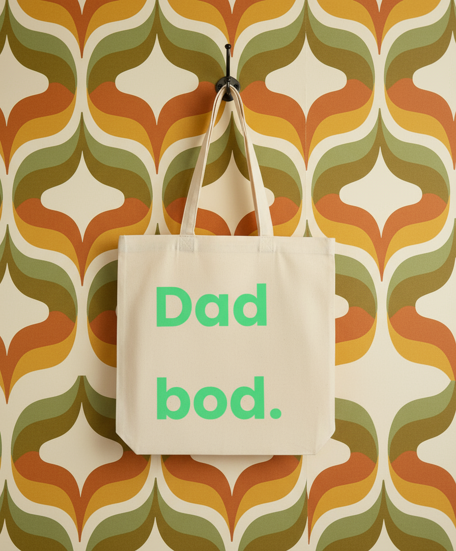 Beige tote bag with 'Dad bod.' text on a retro-patterned wall.