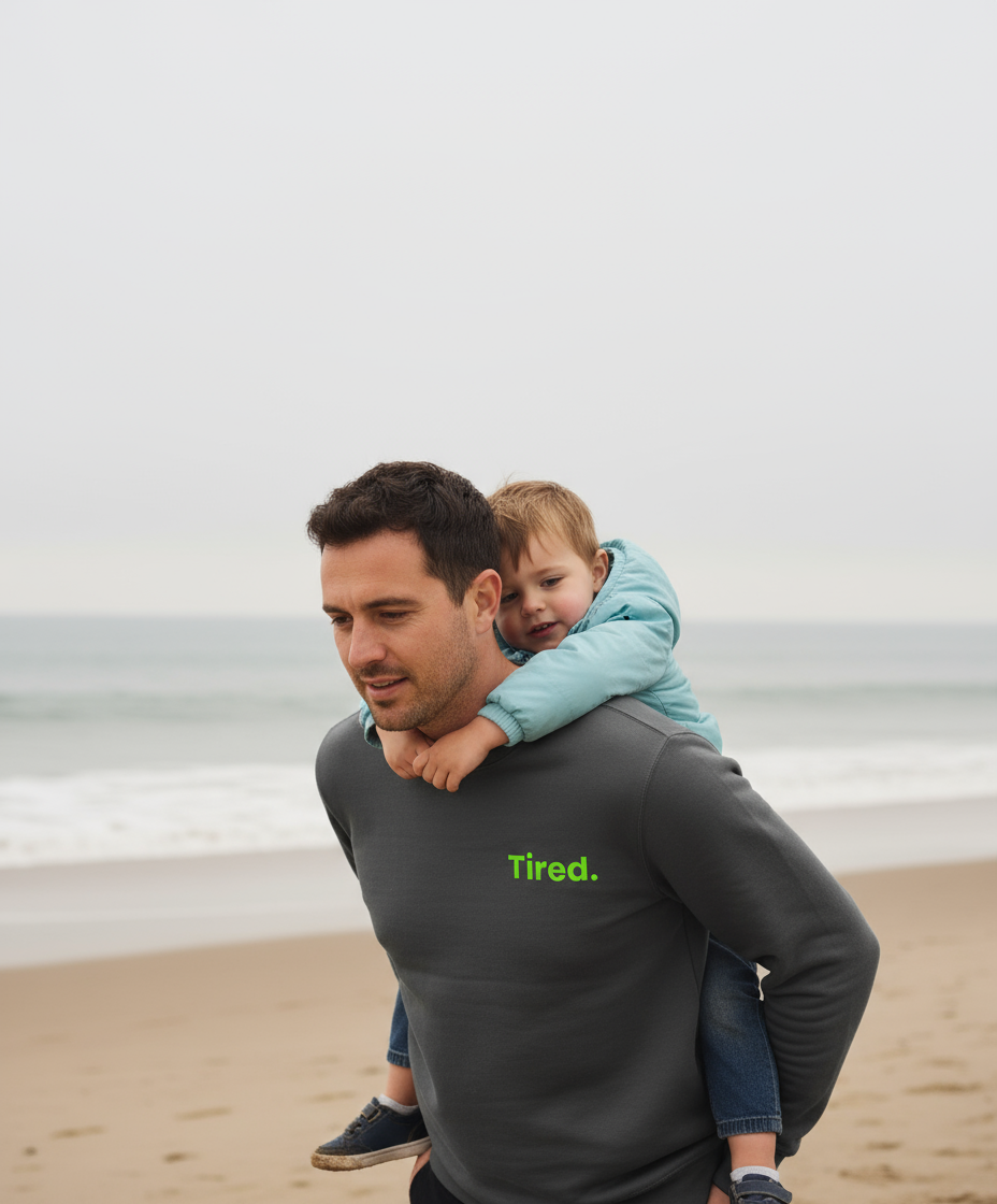 Man carrying a child on his shoulders at the beach with 'Tired.' shirt text.