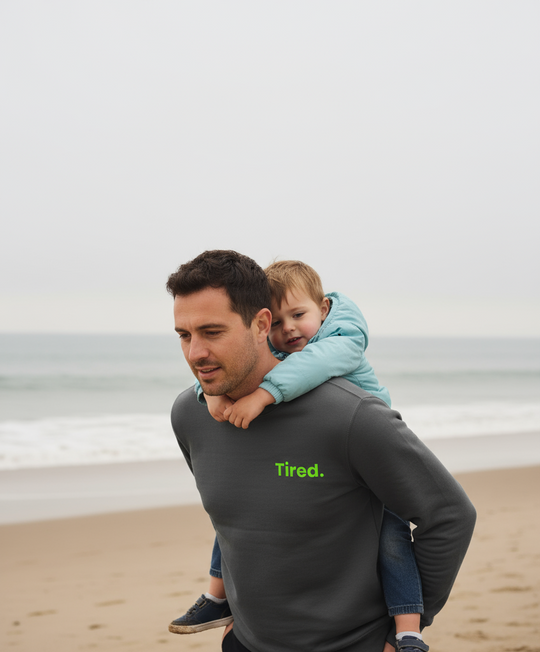 Man carrying a child on his shoulders at the beach with 'Tired.' shirt text.