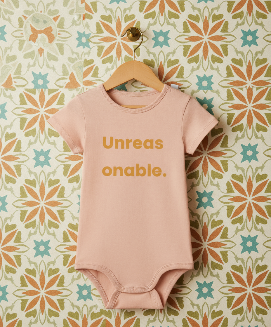 Pink baby onesie with 'Unreasonable' text on a floral patterned wall.
