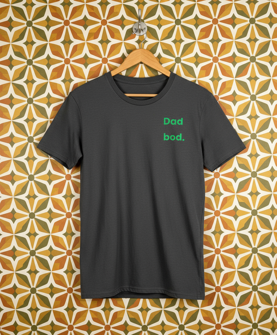 Black t-shirt with 'Dad bod.' text on a hanger against a patterned wall.