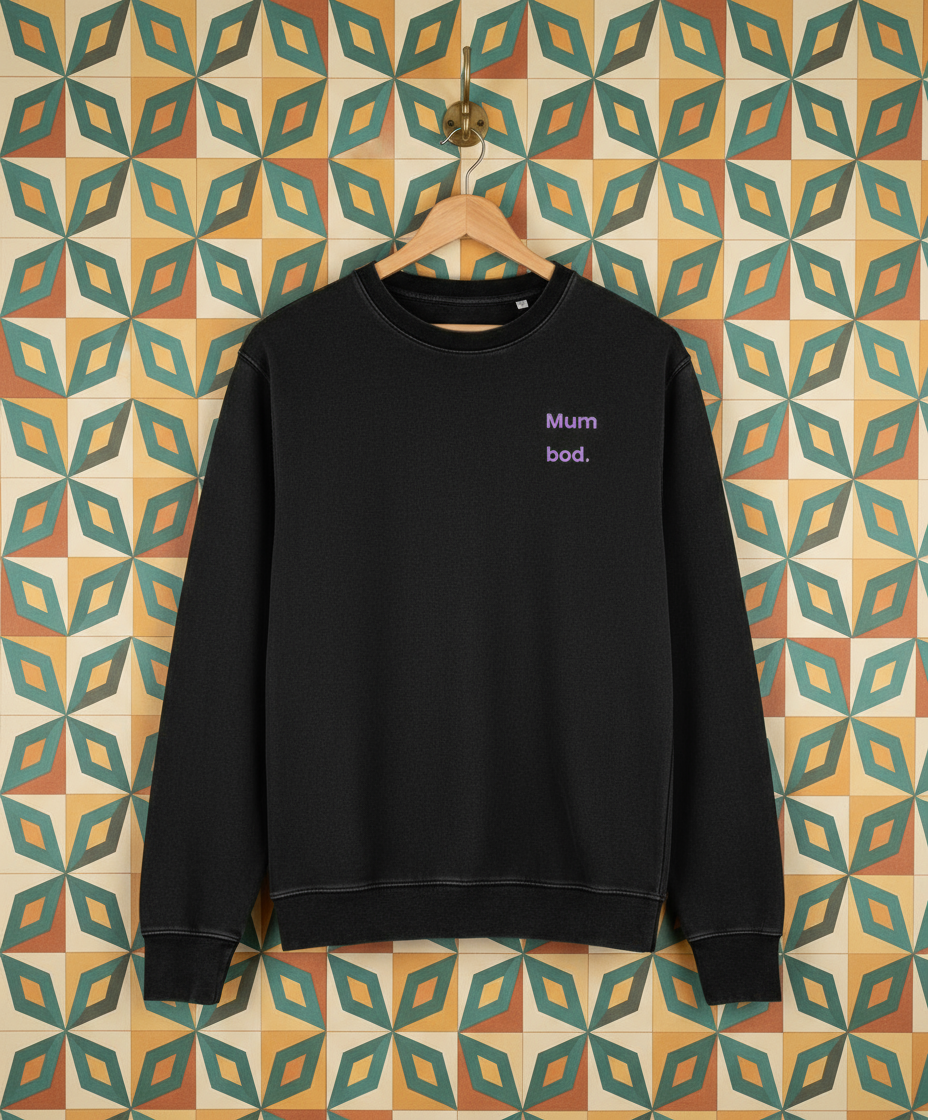 Black sweatshirt with 'mum bod' test hanging on a hook on a wall with retro wallpaper
