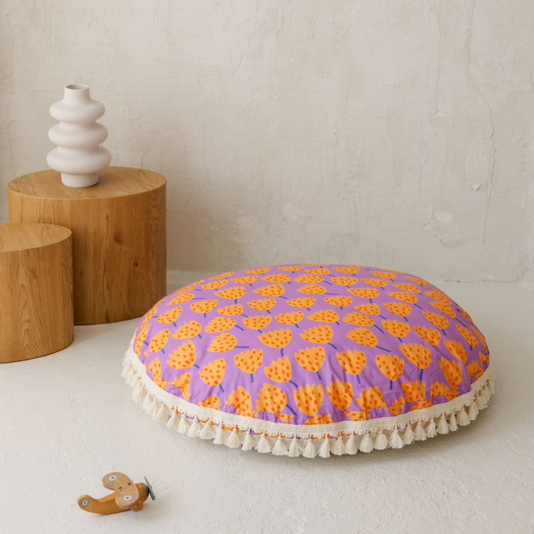 Round purple cushion with orange patterns on a white surface, with wooden stools and a vase in the background.
