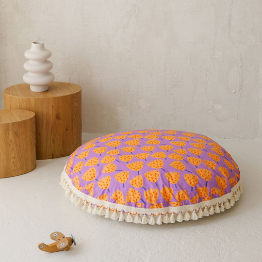 Round purple cushion with orange patterns on a white surface, with wooden stools and a vase in the background.