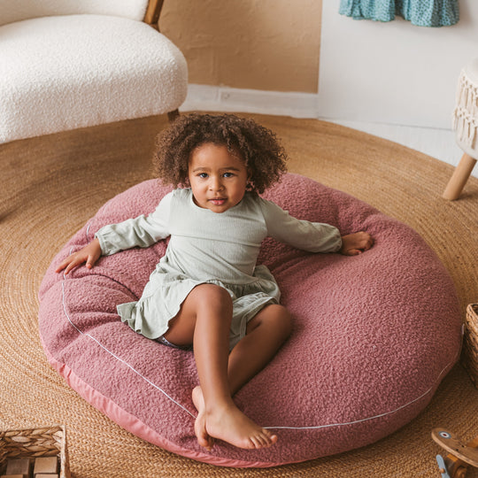 Child sitting on a pink cushion in a cosy room with a textured floor and soft lighting.