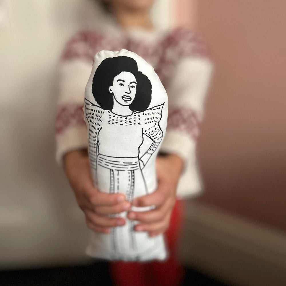 A child holding out a handmade screen printed cushion doll featuring a black and white illustration of Chimamanda Ngozi Adichie towards the camera